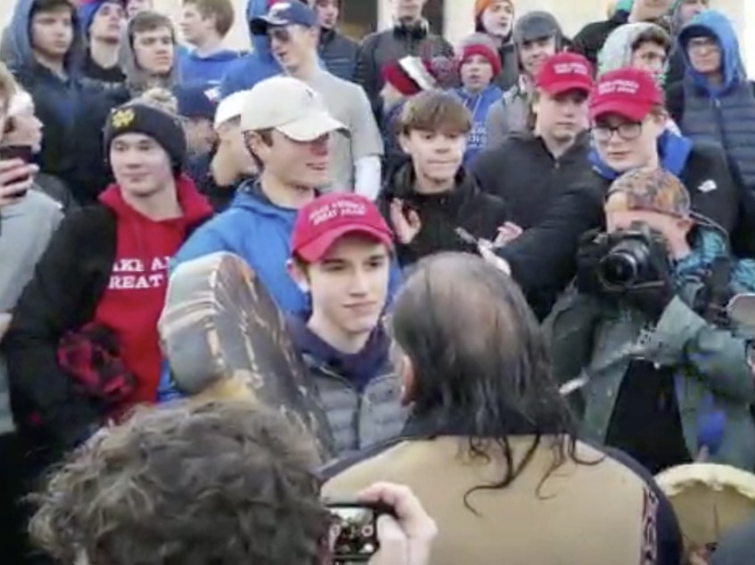 caption: A student wearing a "Make America Great Again" hat stands in front of a Native American singing and playing a drum in Washington on Friday. Young men and women surrounded Nathan Phillips, and a video of the incident has drawn outrage.