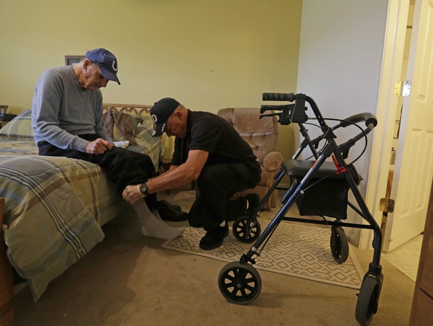 caption: Caregiver Warren Manchess helps Paul Gregoline with his shoes and socks, in Noblesville, Ind. (Darron Cummings/AP)