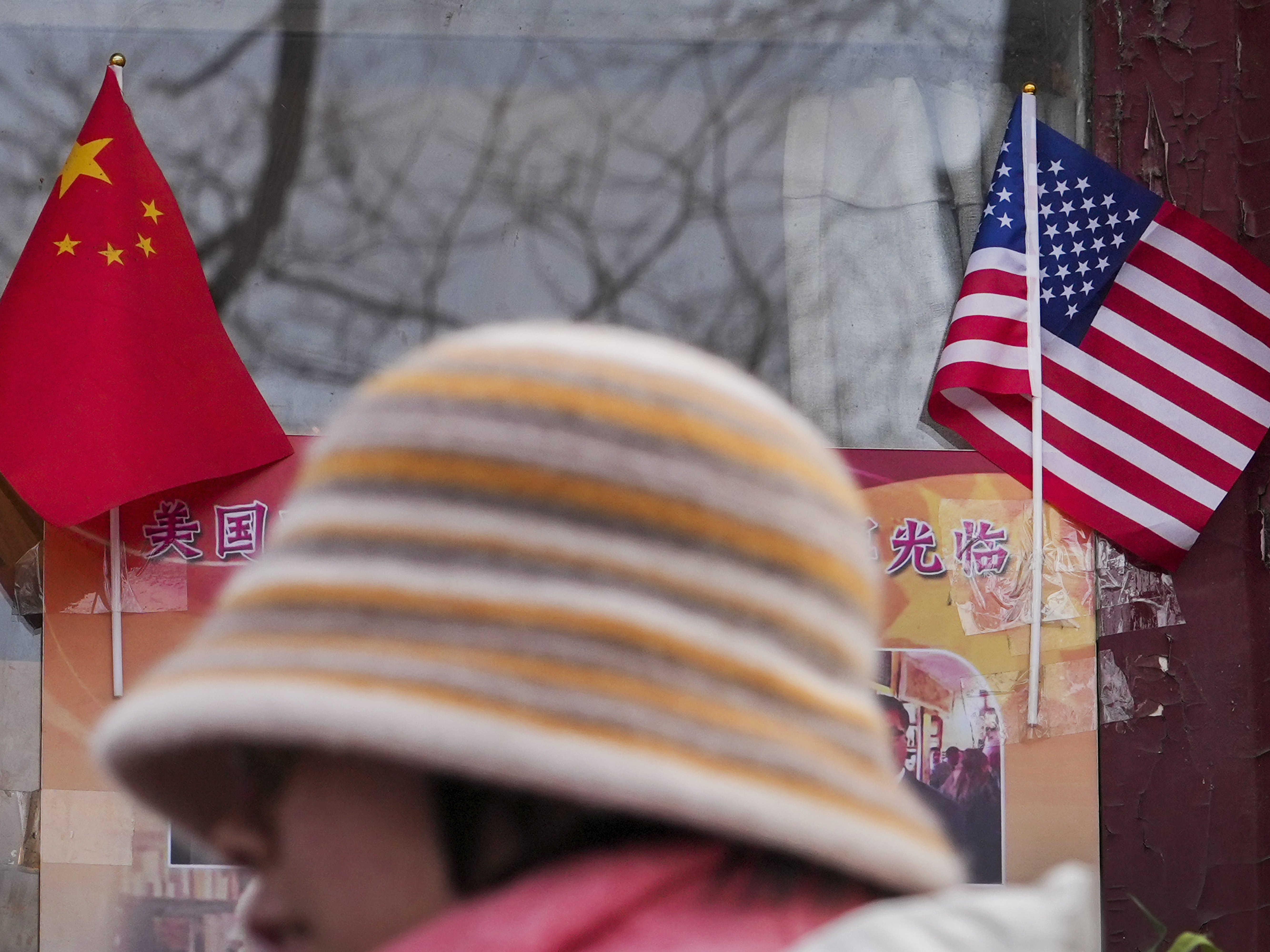 caption: A woman walks by the Chinese and U.S. national flags on display outside a souvenir shop in Beijing on Jan. 31, 2025.
