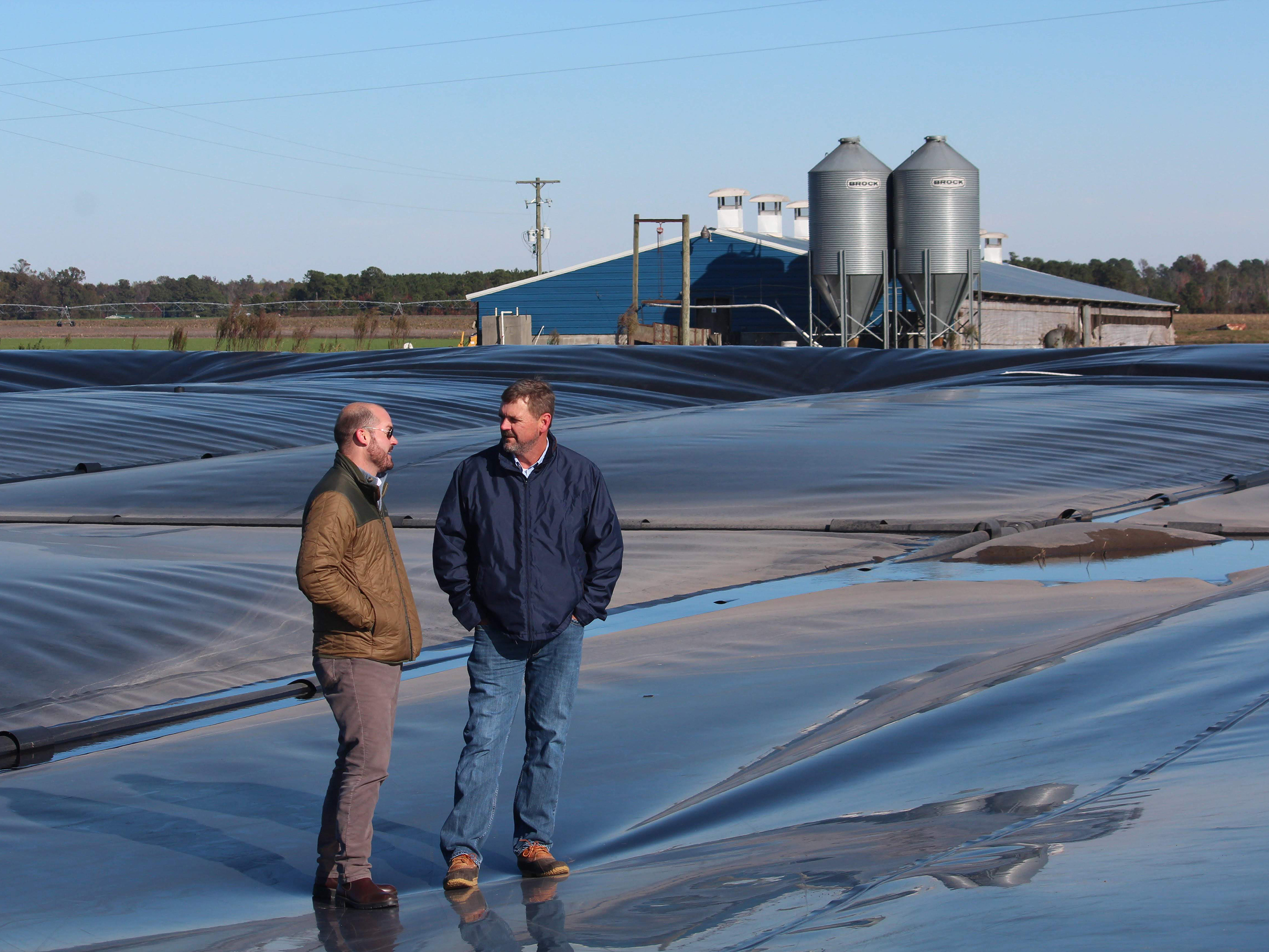 caption: Ryan Childress (left), Dominion Energy's director of gas business development, and Kraig Westerbeek, an executive at pork producer Smithfield Foods, stand on a plastic-covered manure pond in North Carolina.