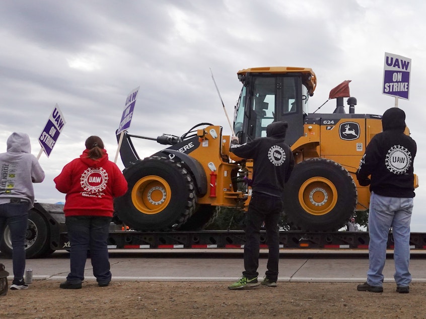 caption: A truck hauls a piece of John Deere equipment from the factory past workers picketing outside of the John Deere Davenport Works facility on Oct. 15, in Davenport, Iowa. More than 10,000 John Deere employees, represented by the UAW, walked off the job in mid-October after failing to agree to the terms of a new contract.