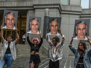 caption: A protest group called "Hot Mess" holds up signs of Jeffrey Epstein on July 8, 2019 in New York City.