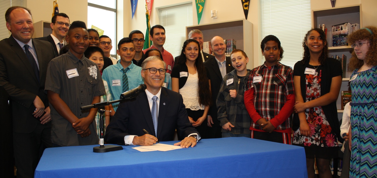 caption: Gov. Jay Inslee signs a bill expanding the College Bound program. Behind him, middle schoolers from Mill Creek Middle School in Kent. At far left is Sen. David Frockt, who wrote the bill.
