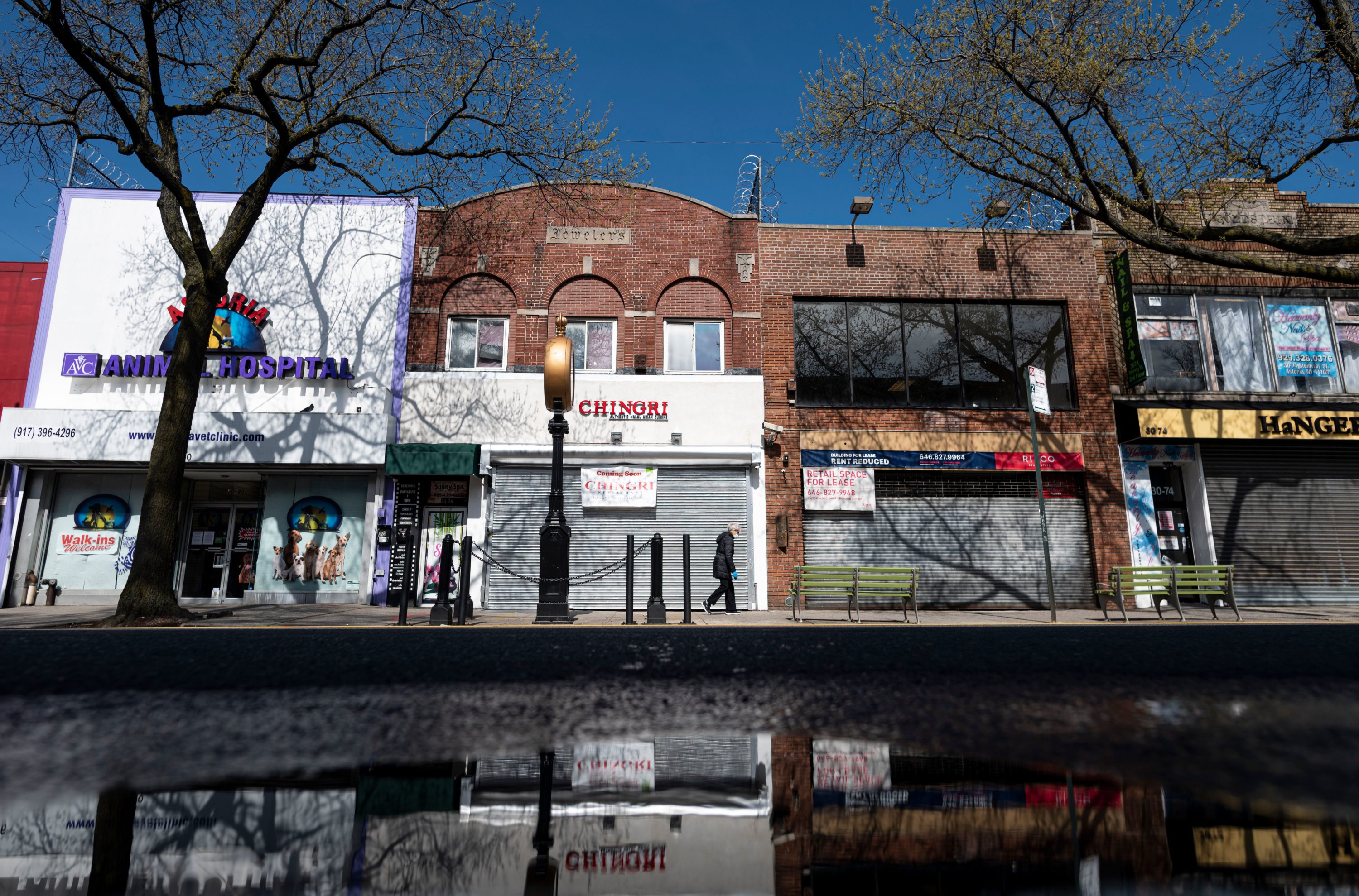caption: A woman wearing a mask walks past closed store fronts in the Astoria neighborhood of Queens, on April 15, 2020 in New York City. (JOHANNES EISELE/AFP via Getty Images)