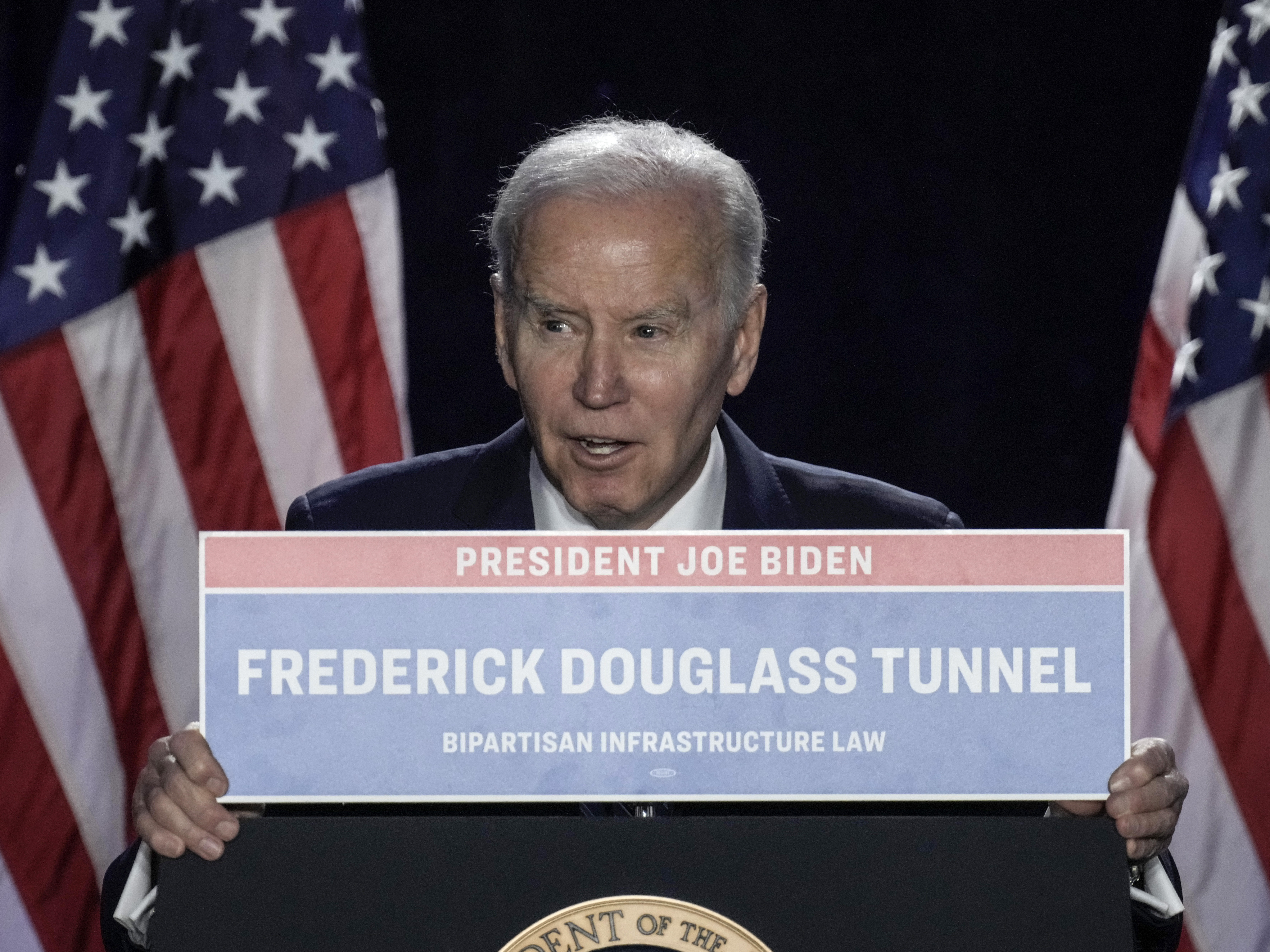 caption: President Biden holds up a sign for the Frederick Douglass Tunnel as he speaks during the annual House Democrats Issues Conference on Wednesday.