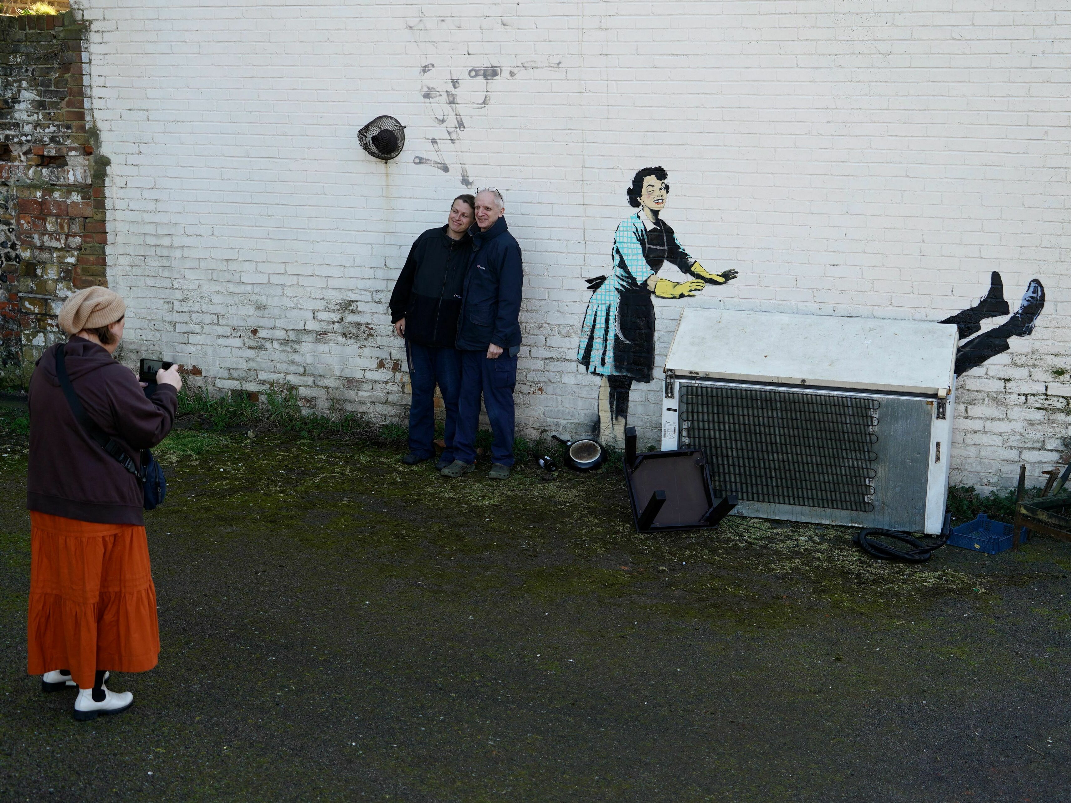 caption: People gathered to see a Banksy artwork on the side of a house in Margate, England, on Valentine's Day. The work depicts a 1950s housewife with a swollen eye, missing a tooth — and apparently shutting a man in a freezer. The freezer was later removed by council workers.