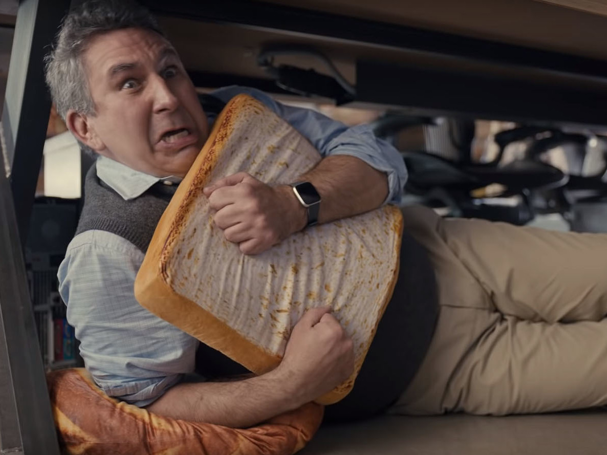 A man cowers under a desk, hugging a pillow shaped like a slice of bread, in a scene from a  Little Caesars commercial that ran during the 2020 Super Bowl.