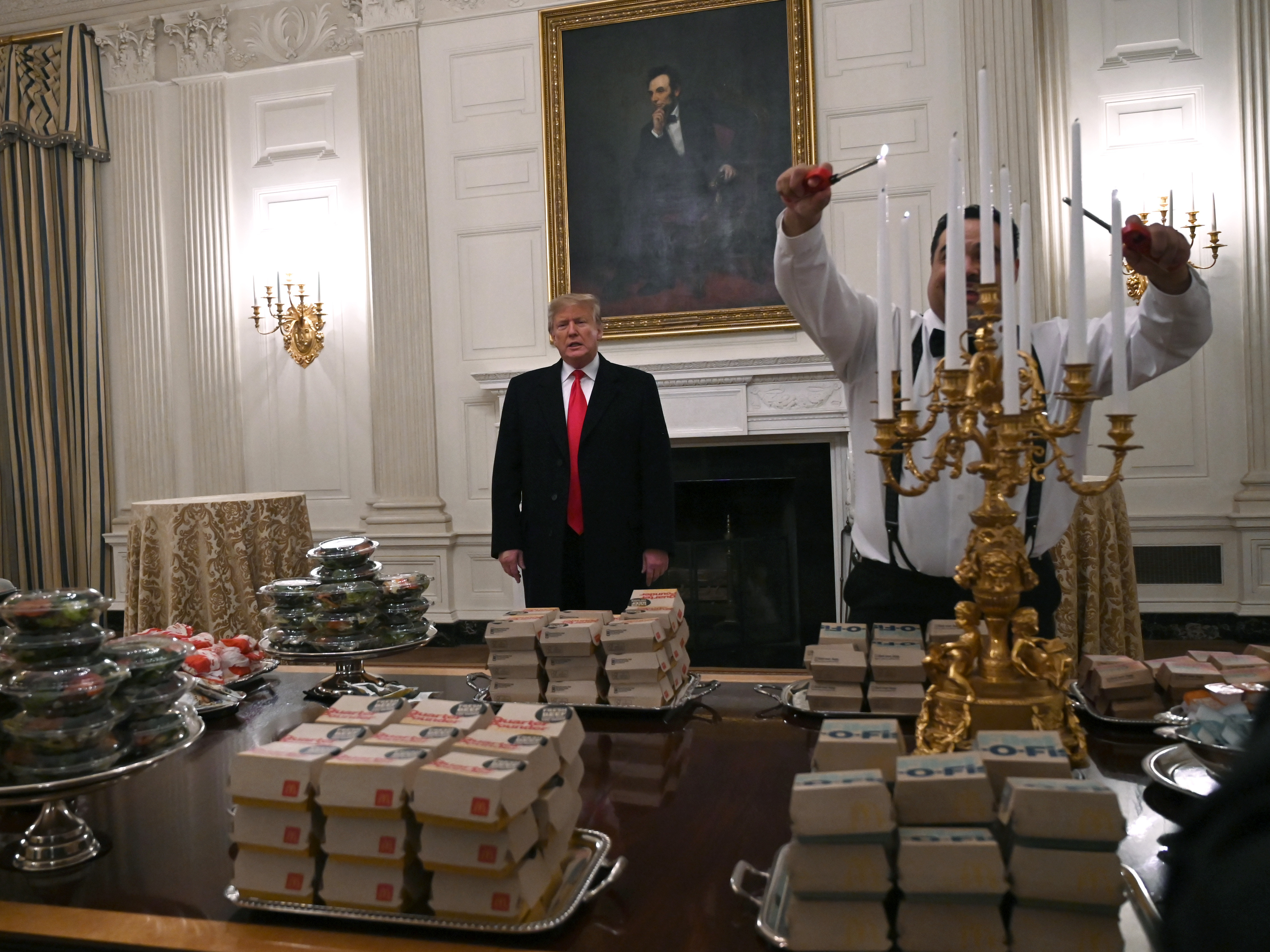 caption: President Trump talks to the press about the table full of fast food laid out in the State Dining Room of the White House for a reception for the Clemson Tigers.