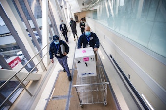 caption: Richard Guarino, Boston Medical Center supply chain operations associate director, wheels a cart containing Pfizer's COVID-19 vaccine to the pharmacy. (Jesse Costa/WBUR)