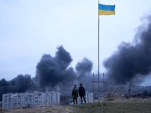 caption: People standing near a Ukrainian national flag watch as dark smoke billows following an air strike in the western Ukrainian city of Lviv, on March 26, 2022.