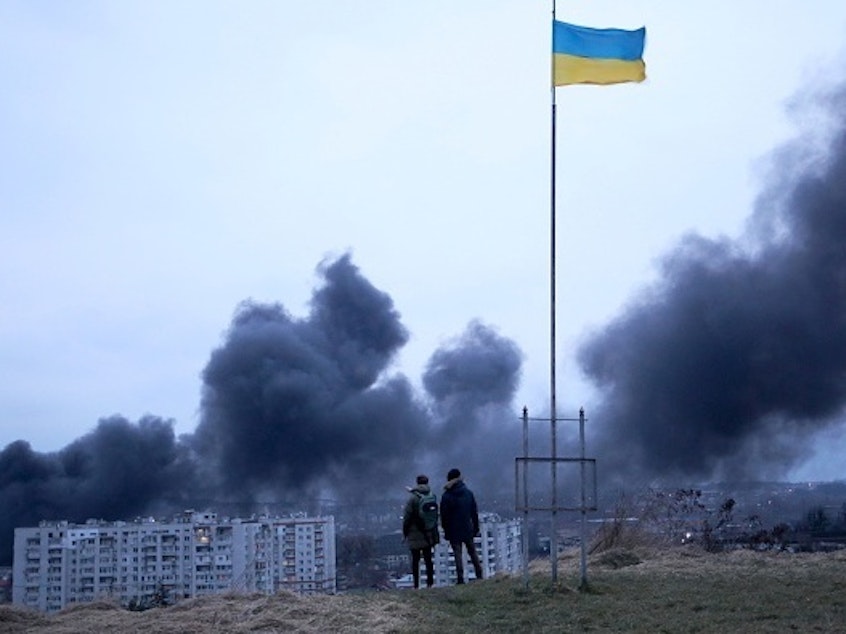 caption: People standing near a Ukrainian national flag watch as dark smoke billows following an air strike in the western Ukrainian city of Lviv, on March 26, 2022.