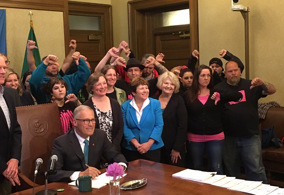 caption: Vape shop owners give the thumbs down sign as Gov. Jay Inslee poses for a photo after signing into law a new tax on vaping liquids.