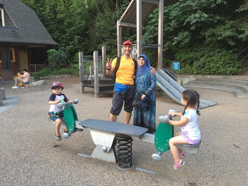 caption: Mariam Khazal's parents Mohammed Khazal and Rasha Ibrahim pose for a photo at a Seattle park while Mariam's little sister and a friend play on the seesaw, summer 2019.