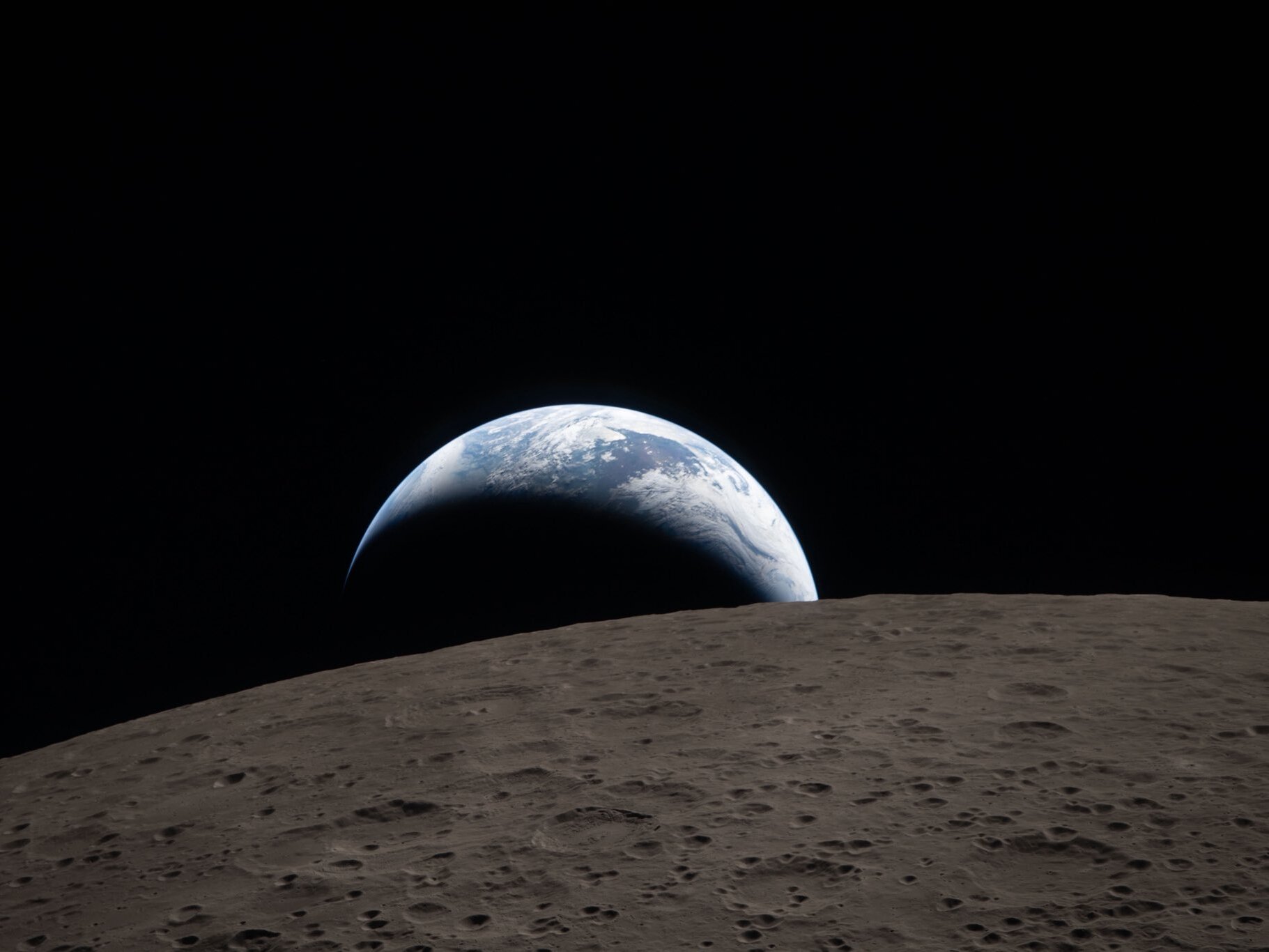 caption: Earthset captured through the Orion spacecraft window at 6:41 p.m. ET on April 6 during the Artemis II crew's flyby of the moon. A muted blue Earth with bright white clouds sets behind the cratered lunar surface. The dark portion of Earth is experiencing nighttime. In the foreground, Ohm crater has terraced edges and a flat floor interrupted by central peaks. Central peaks form in complex craters when the lunar surface, liquefied on impact, splashes upward during the crater's formation.