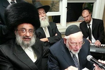 caption: The late Hasidic composer Ben Zion Shenker sings, wearing a yarmulke and holding a mic, at a male-only sing along known as a k<em>umzits </em>that took place in an Orthodox neighborhood in Brooklyn.