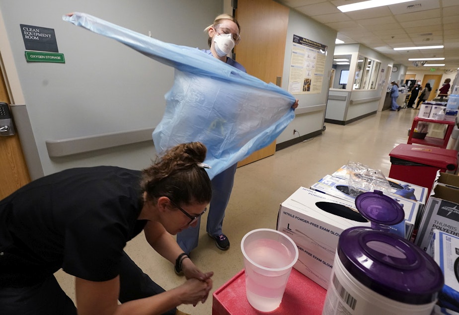 caption: Registered nurse Jessalynn Dest, left, removes protective equipment and washes her hands after leaving a COVID-19 patient's room as speech therapist Sam Gibbs puts on safety clothing while preparing to see a patient in the acute care unit of Harborview Medical Center, Friday, Jan. 14, 2022, in Seattle.
