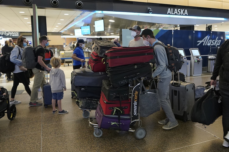caption: Passengers wearing masks walk with luggage near an Alaska Airlines check-in area, Monday, March 1, 2021, at Seattle-Tacoma International Airport in Seattle. 