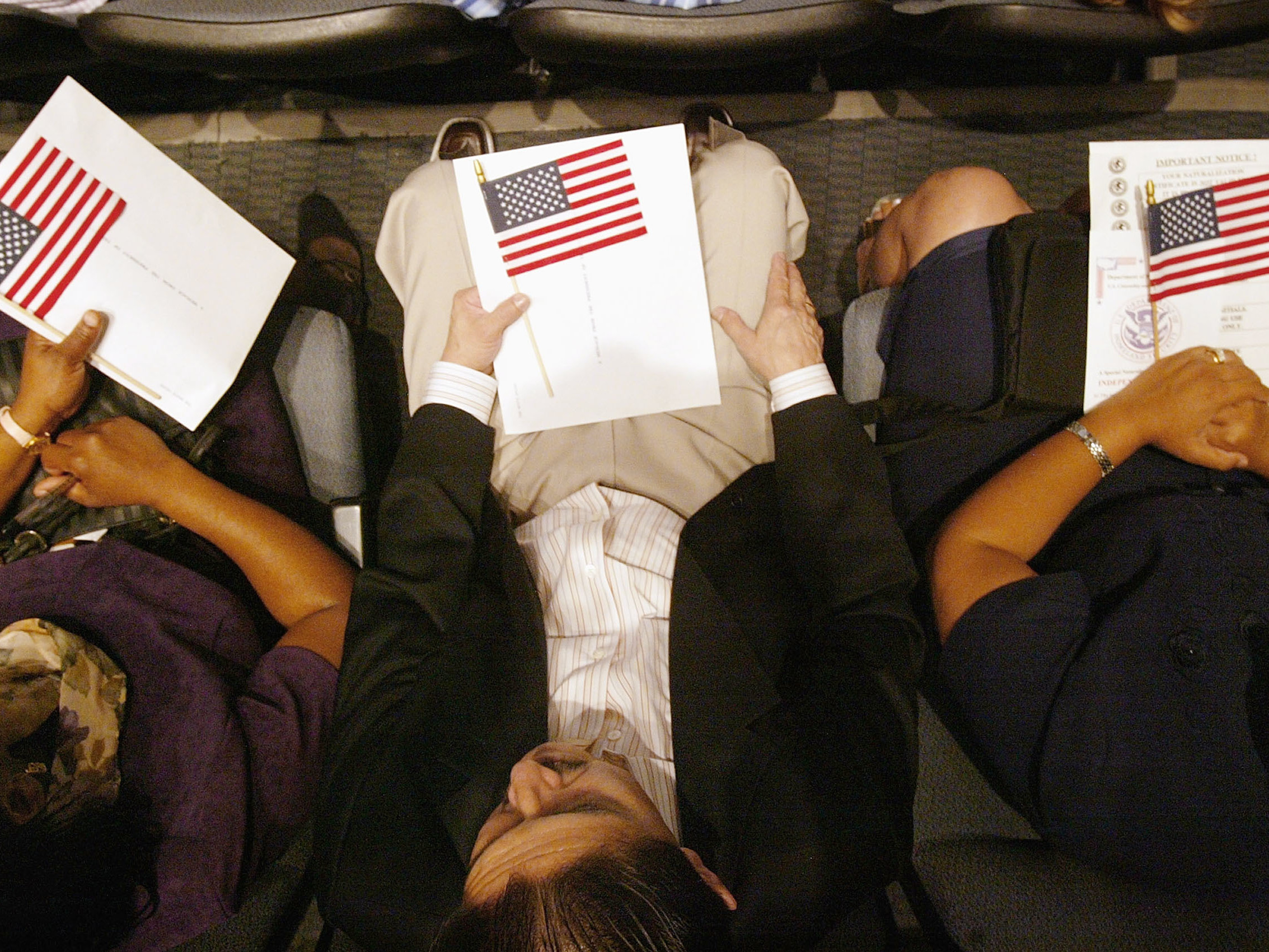caption: Three people hold U.S. flags as they wait to be sworn in as American citizens at a naturalization ceremony on the Intrepid Sea-Air-Space Museum.