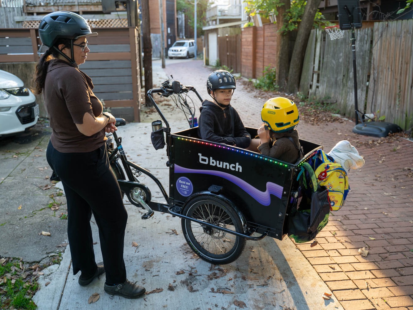 caption: Lelac Almagor waits as her kids get ready for the ride to school. Her cargo bike is technically a trike, with two wheels in front for more stability.