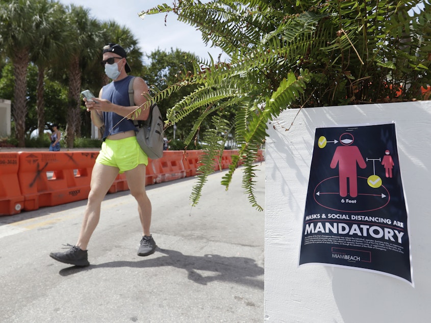 caption: A pedestrian in a mask passes a sign urging people to practice social distancing, on Saturday in Miami Beach, Fla. Just as residents flocked outside to enjoy the Fourth of July, states such as Florida were reporting skyrocketing numbers of confirmed coronavirus cases.