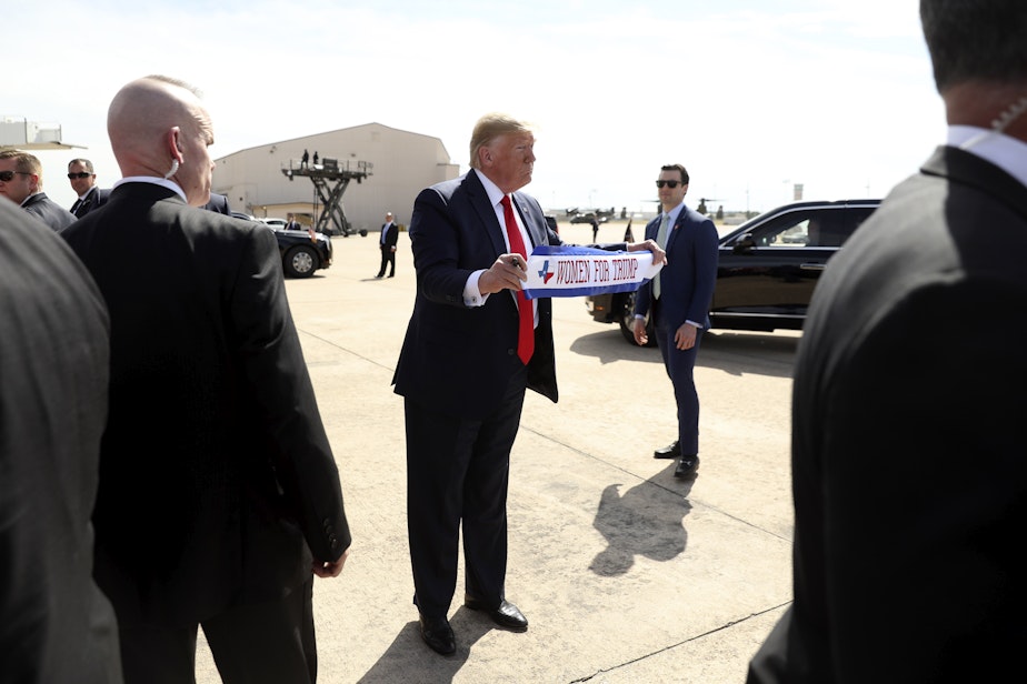 caption: President Donald Trump holds a banner, "Women for Trump," as he talks with guests as he arrives at Naval Air Station Joint Reserve Base in Fort Worth, Texas, Thursday, Oct. 17, 2019. 