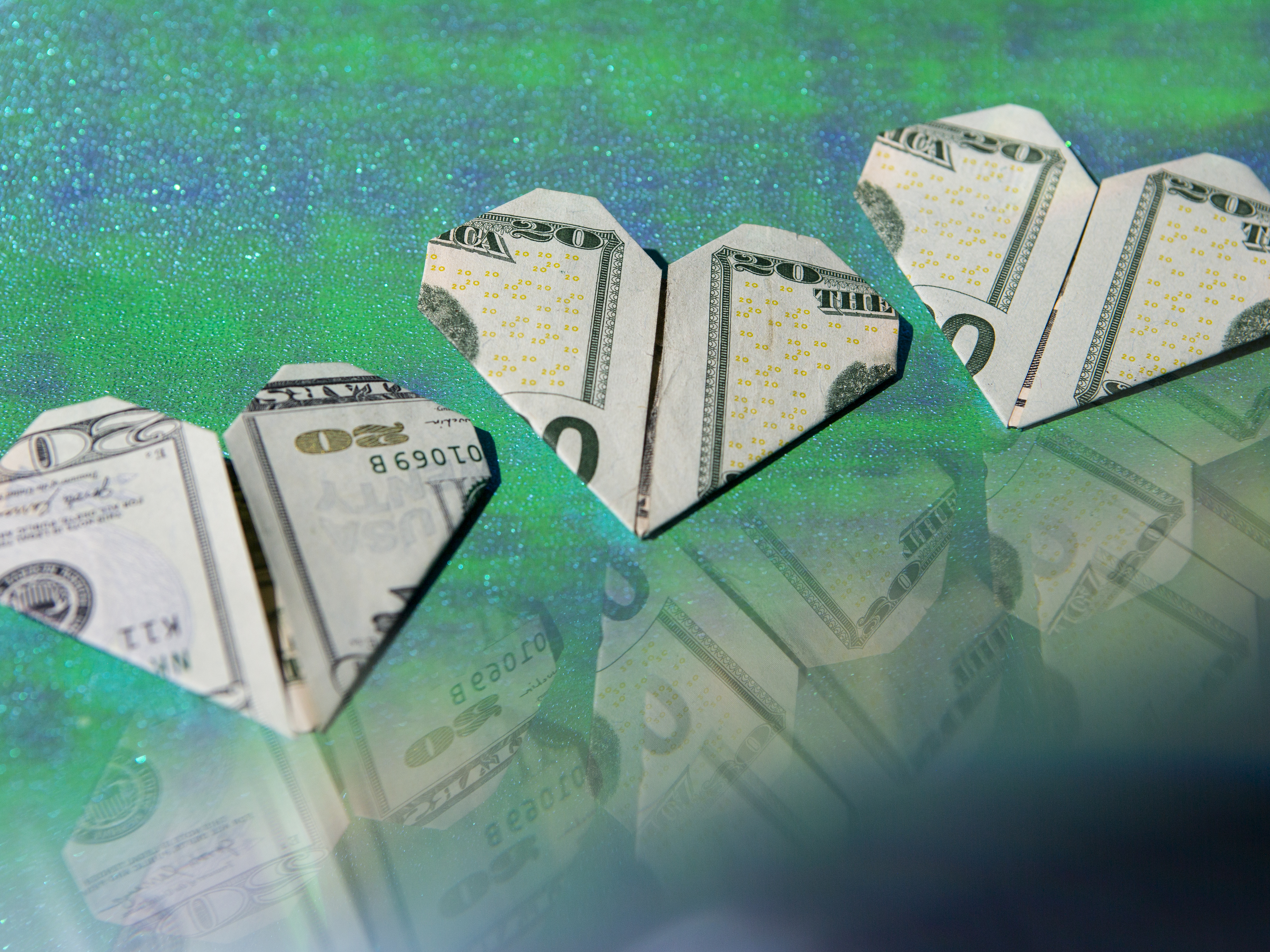Three 20 dollar bills folded into hearts are arranged in an upward trending diagonal line on a sparkly green background, reflections of the money are cast to the lower right side of the frame.