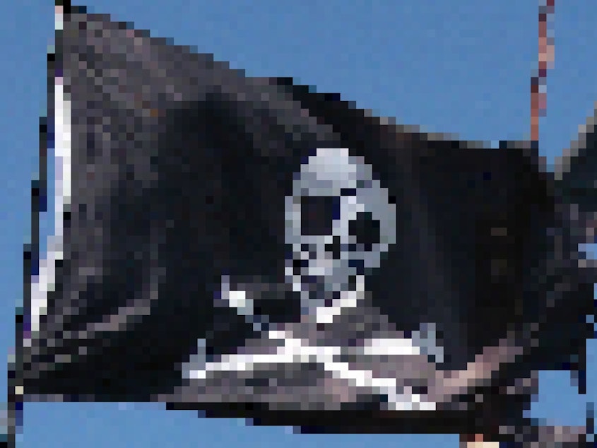 A classic pirate flag with white skull and crossbones on a black background. It's torn on the edges and flying against a bright blue sky.