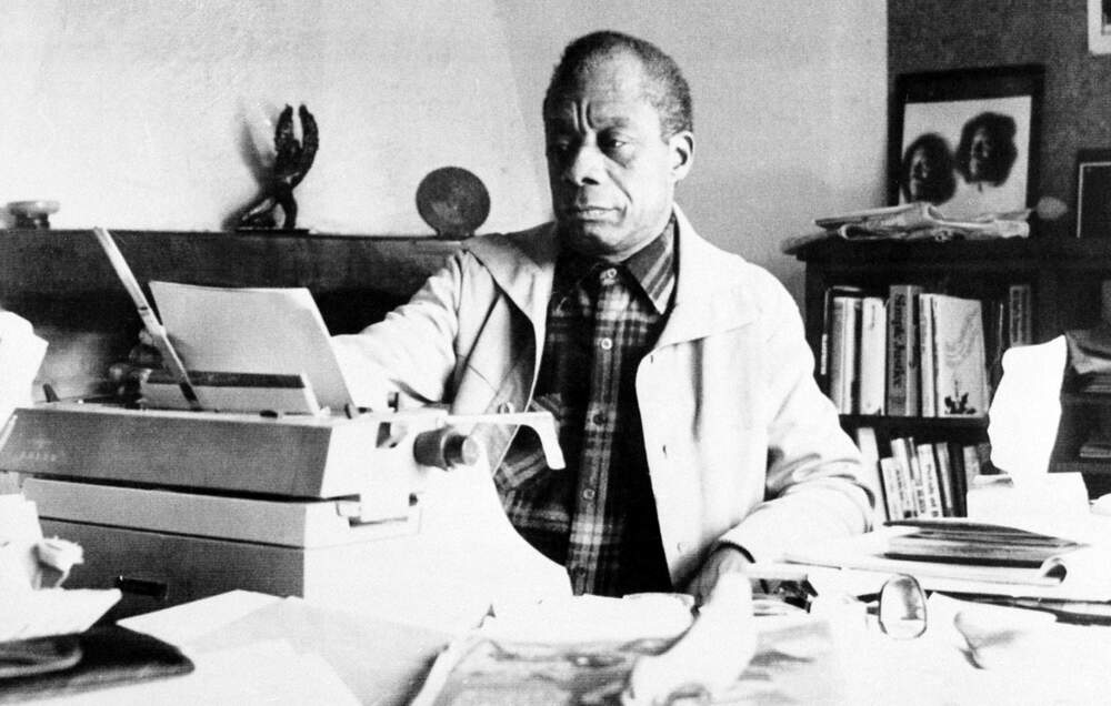 caption: American novelist James Baldwin sits in front of his typewriter in the study of his home in St. Paul de Vence, in southern France, on March 21, 1983.  (AP Photo)
