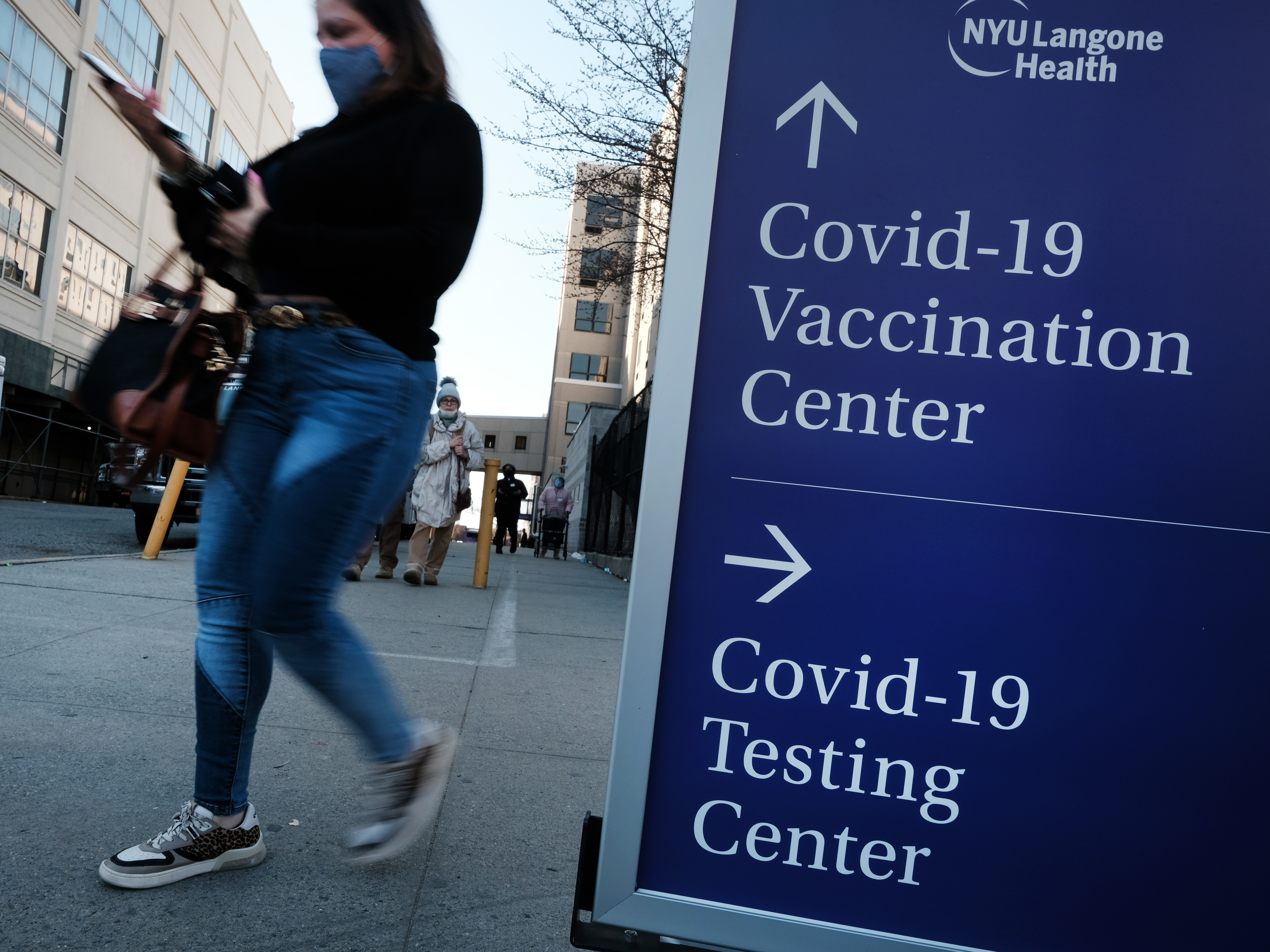 caption: People walk by a sign for both a COVID-19 testing clinic and a Covid vaccination location outside of a Brooklyn, New York, hospital on March, 29 2021.