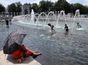 caption: Visitors and tourists to the World War II Memorial in Washington, D.C., seek relief from the hot weather in the memorial's fountain on July 3. Due to extreme temperatures and high humidity, D.C. has declared a heat emergency, urging residents to take precautions outside and to stay hydrated.