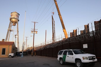 caption: A U.S. Border Patrol vehicle is stationed in front of the U.S.-Mexico border barrier as construction continues in hard-hit Imperial County on July 22, in Calexico, Calif.