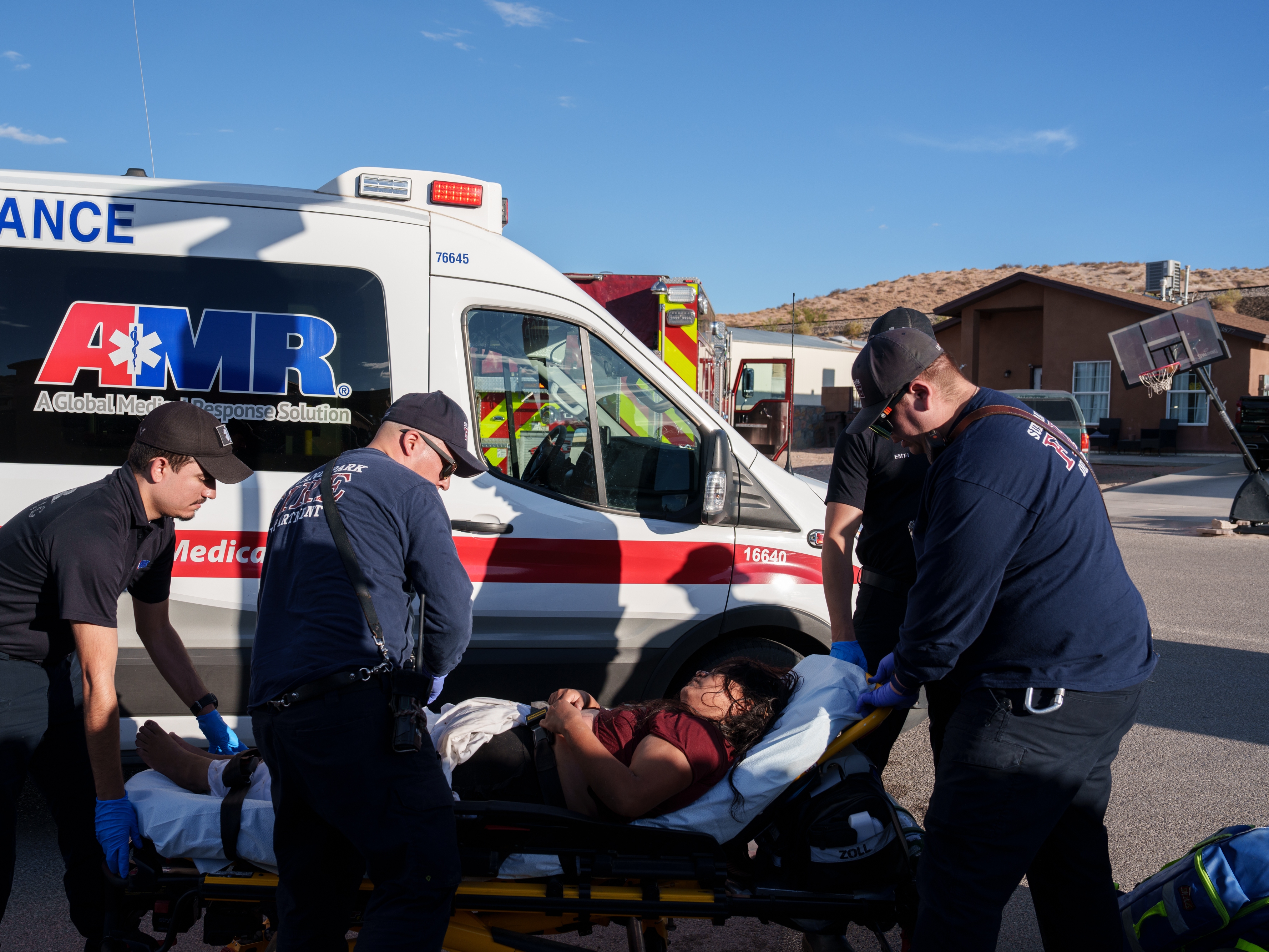 caption: Sunland Park Fire Department firefighters and police officers roll a migrant woman, who was suffering from heat-related illness, on a gurney to be loaded into an ambulance in a residential area in Sunland Park, N.M., on Friday, June 28, 2024.