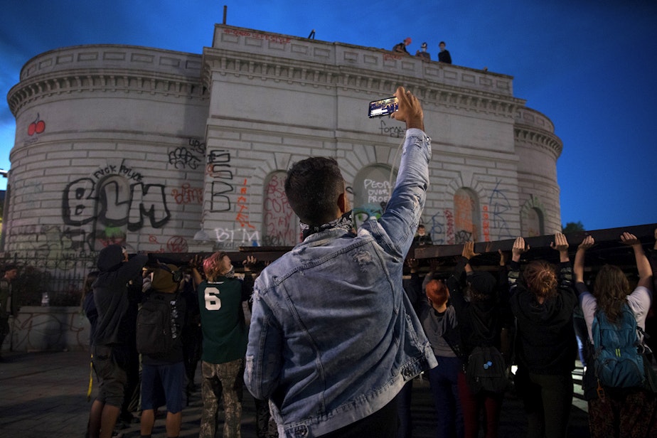 caption: A protester live-streams as others attempt to lift a Black Power fist art instillation inside the Capitol Hill Occupied Protest Zone on Tuesday, June 16, 2020, in Seattle. As a result of safety concerns, it was decided that the structure would be raised the following day instead.
