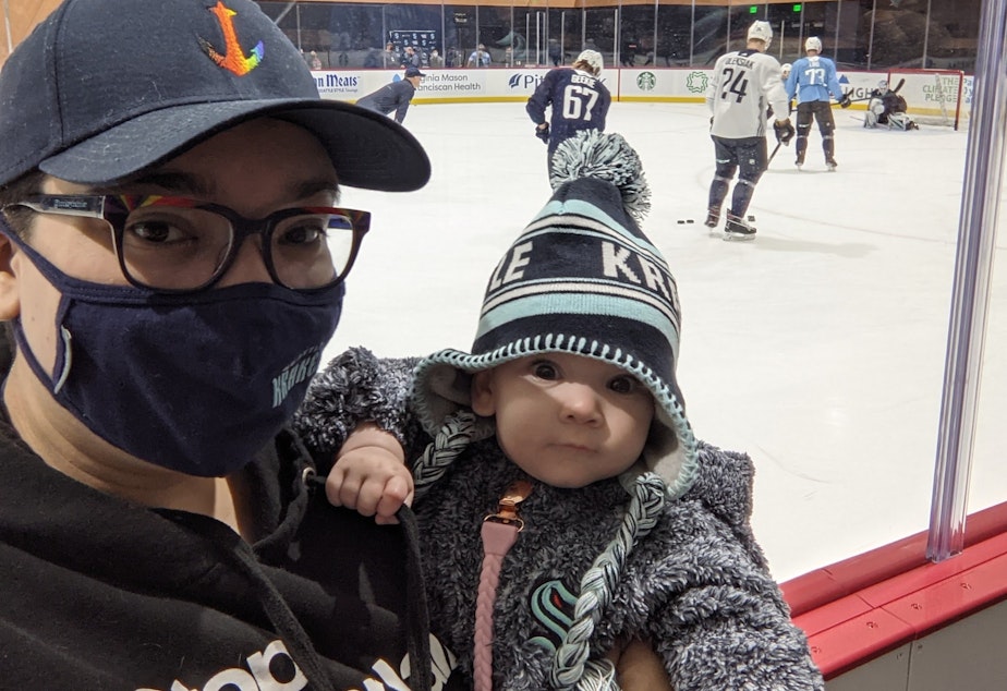 caption: Christy Maggio, a mega hockey fan, with their baby. Maggio loves the Kraken, Seattle's new hockey team, but laments misogyny heard from the stands.