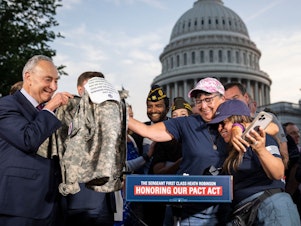 caption: Senate Majority Leader Chuck Schumer (D-N.Y.) looks on Tuesday as Susan Zeier, mother-in-law of the late Sgt. First Class Heath Robinson, hugs Rosie Torres, wife of veteran Le Roy Torres, who suffers from illnesses related to his exposure to burn pits in Iraq, after the Senate passed the PACT Act at the U.S. Capitol in Washington.