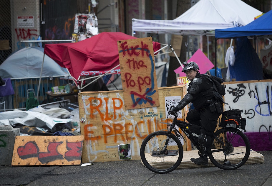 caption: A Seattle Police Department officer rides a bicycle next to a 'Fuck the PoPo' sign after the Capitol Hill Organized Protest zone was cleared by Seattle Police Department officers early Wednesday morning, July 1, 2020, at the intersection of 12th Avenue and East Pine Street in Seattle. 
