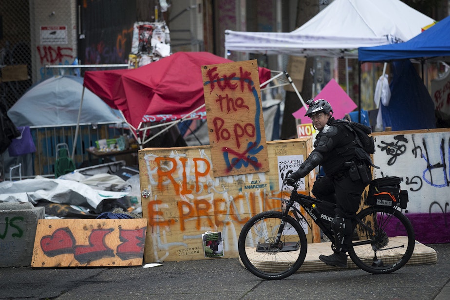 caption: A Seattle Police Department officer rides a bicycle next to a 'Fuck the PoPo' sign after the Capitol Hill Organized Protest zone was cleared by Seattle Police Department officers early Wednesday morning, July 1, 2020, at the intersection of 12th Avenue and East Pine Street in Seattle. 