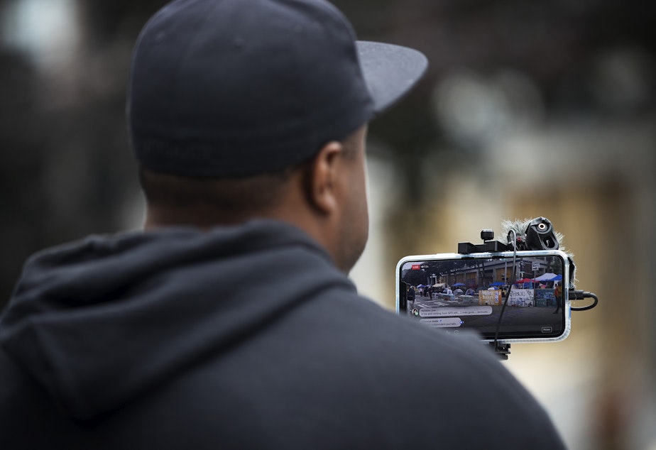 caption: Omari Salisbury, a journalist with Converge Media, live-streams at the intersection of 12th and Pine Streets on Monday, June 29, 2020, inside the Capitol Hill Organized Protest zone in Seattle. Earlier that morning, a 16-year-old boy was killed and a 14-year-old boy was critically injured in a shooting.