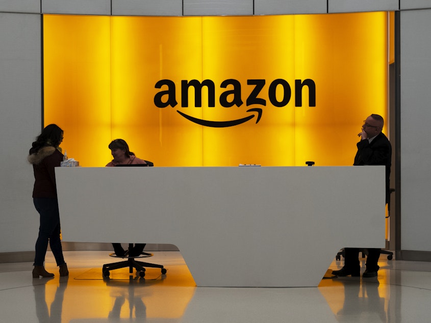 caption: People stand in the lobby of Amazon offices in New York in January 2019. At a time of mass work from home and many moving to spacious suburbs, Amazon is funding a large expansion of corporate real estate and jobs in New York and five other U.S. cities.