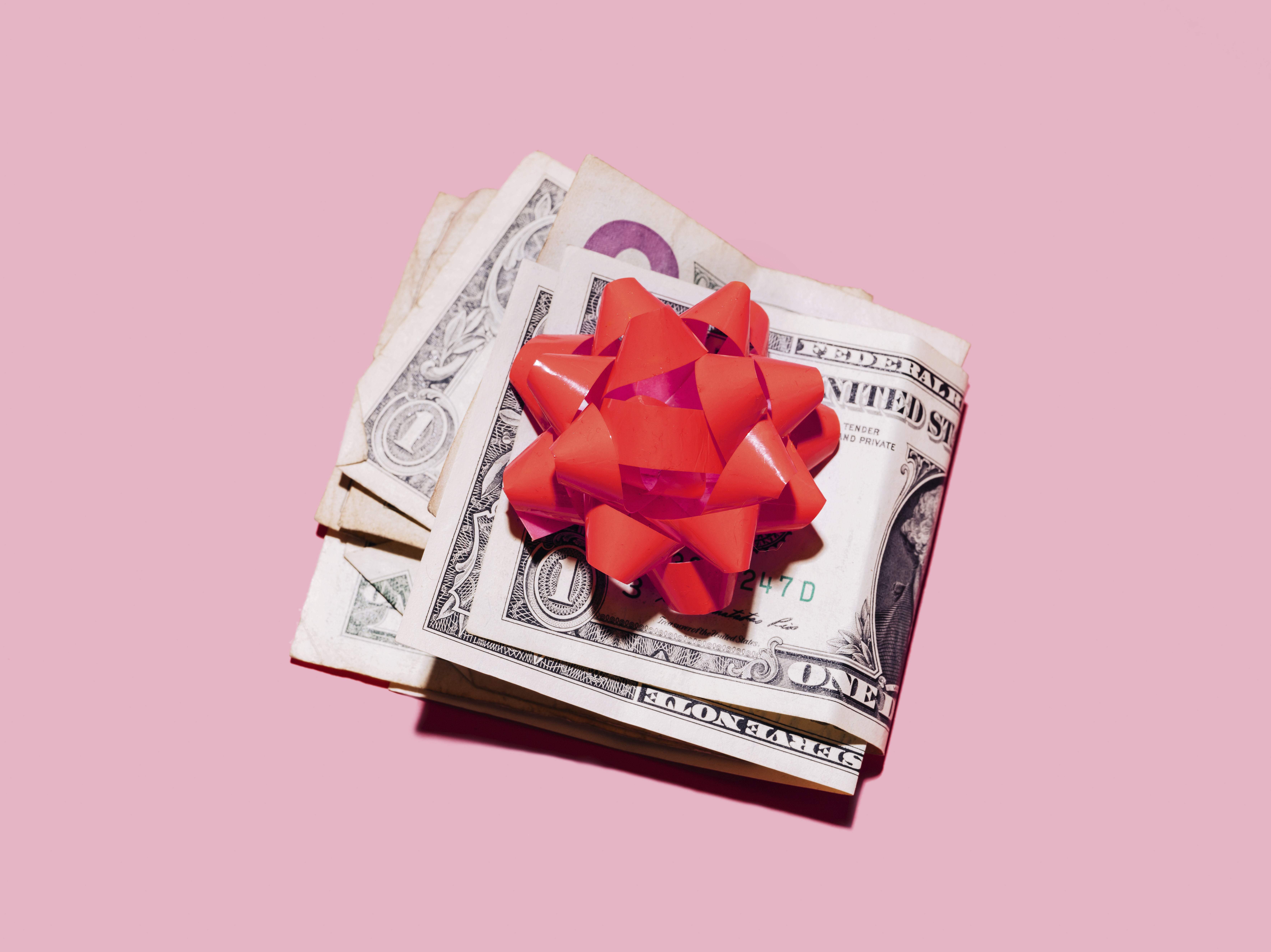 Photo of folded dollar bills with a red gift ribbon placed on top against a light pink background.