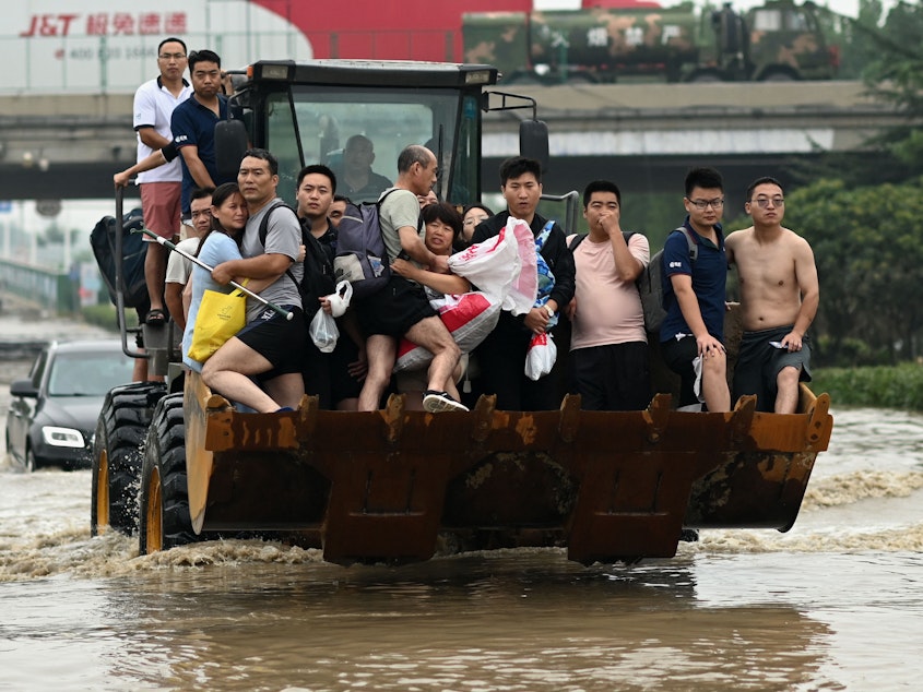 caption: People ride in the front of a wheel loader to cross a flooded street following heavy rains which caused flooding and claimed the lives of at least 63 people in the city of Zhengzhou in China's Henan province on July 23.