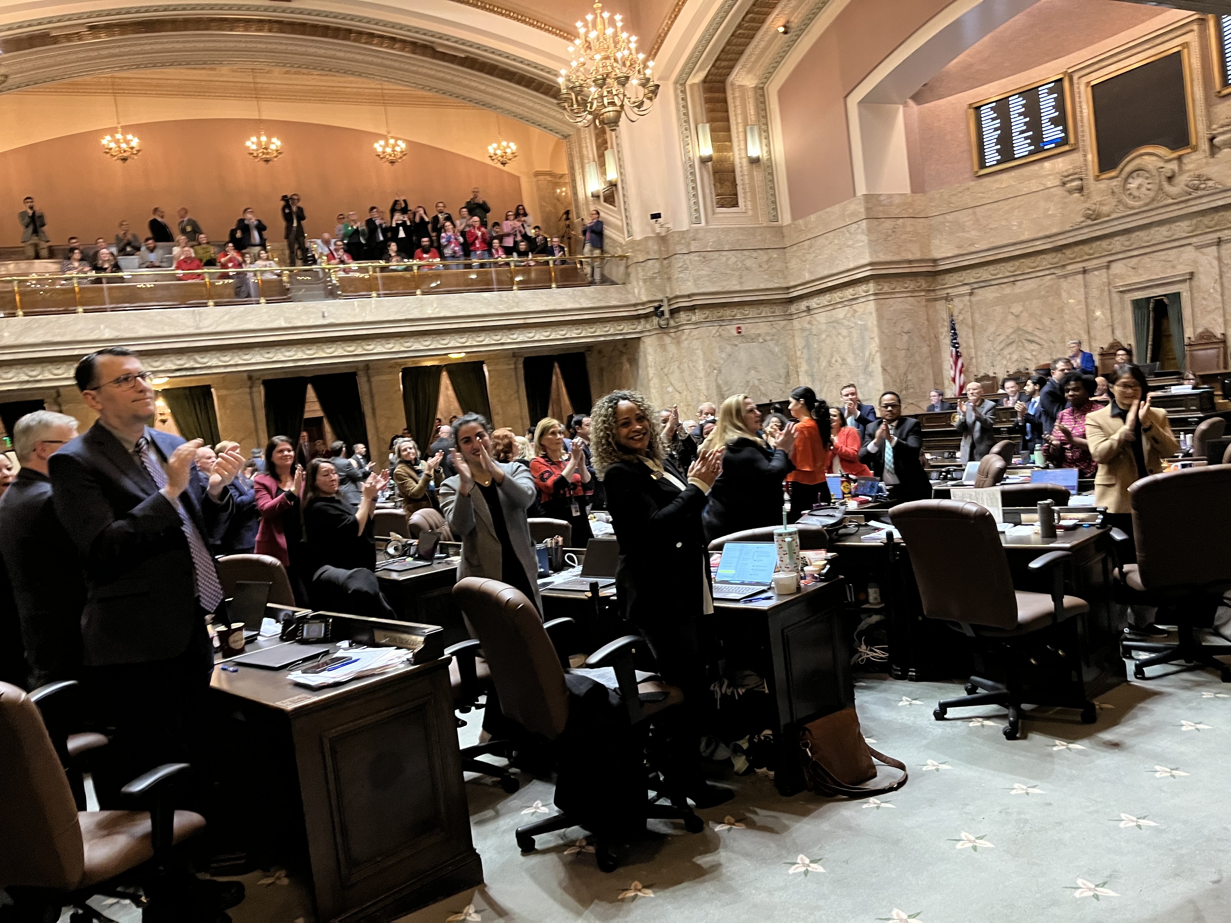 caption: Washington state House representatives stand and clap after a final vote was tallied on the proposed income tax on high earners, also known as the "millionaires' tax," on March 10, 2026, at the state Capitol in Olympia.