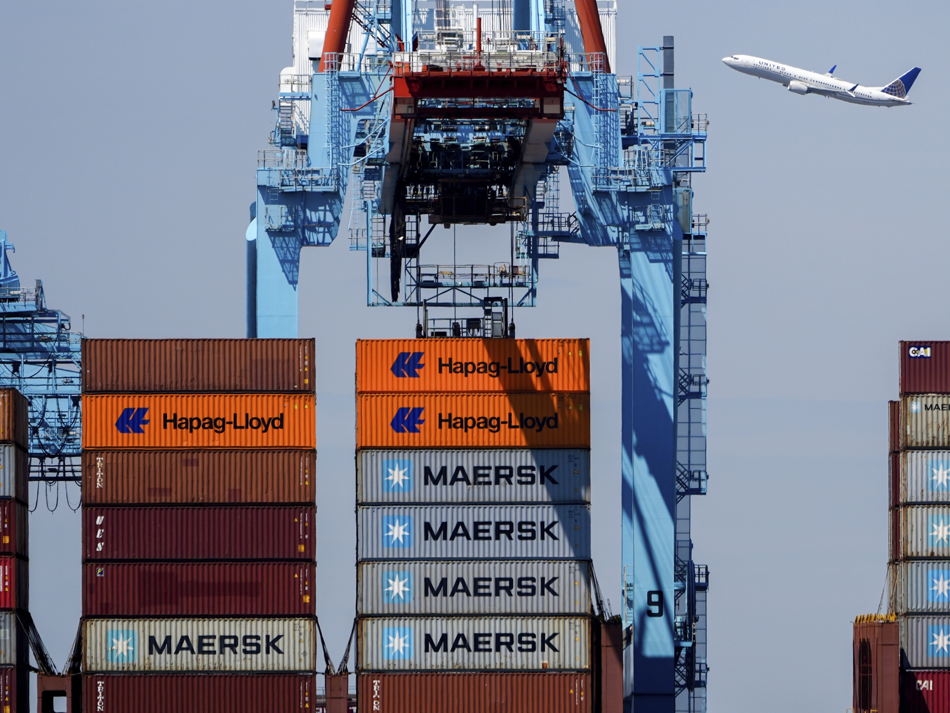 caption: An airplane flies over the container ship Alexandra at the Port Newark Container Terminal on April 18 in Newark, N.J.