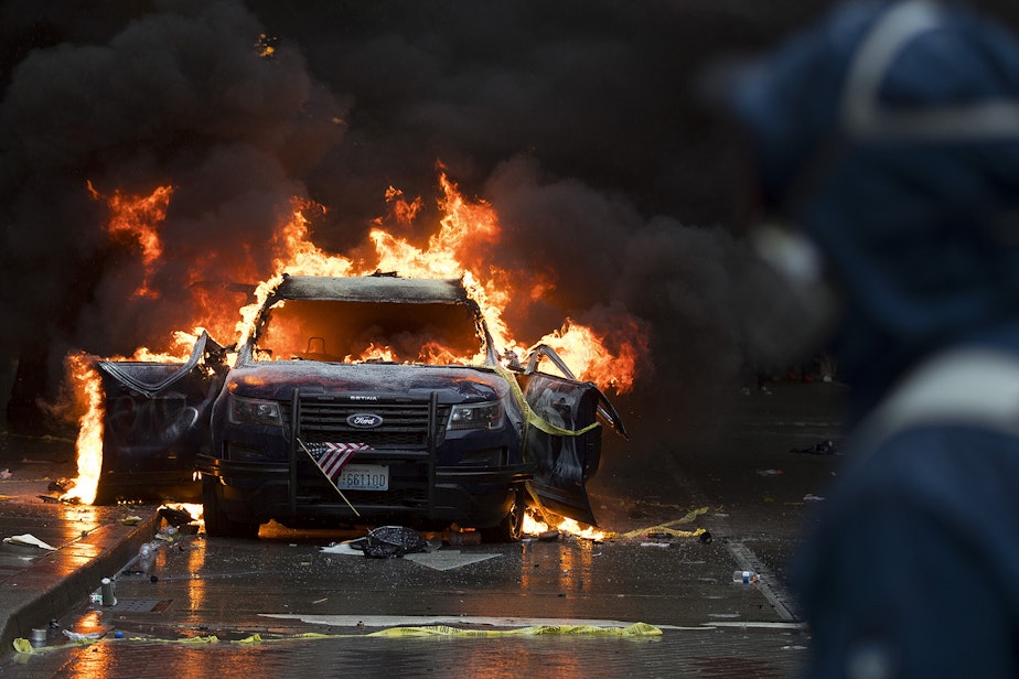 caption: A protester wearing a respirator watches as a small American flag hangs from the grill of a burning Seattle Police Department cruiser as thousands gathered in protest following the murder of George Floyd on Saturday, May 30, 2020, near the intersection of 5th Avenue and Pine Street in Seattle.