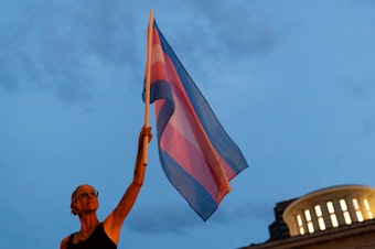 caption: Cole Ramsey, 39, of South Linden, Ohio, holds a Transgender Pride Flag in front of the Ohio Statehouse in Columbus to protest the passing of legislation against trans women playing sports in high school and college.