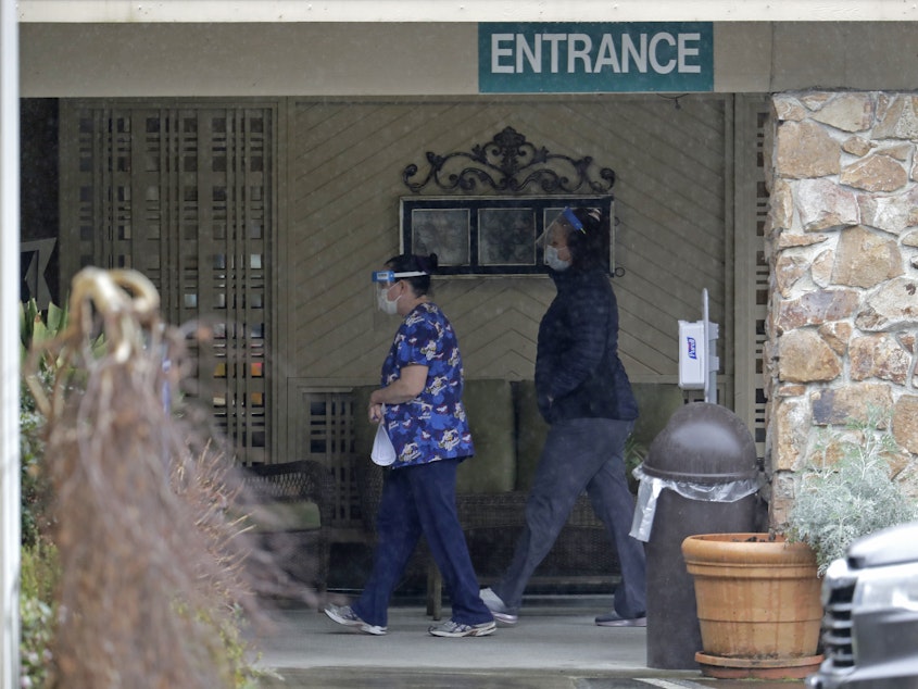 caption: Two workers approach the entrance to Life Care Center in Kirkland, Wash., on March 13. An association that represents nursing homes is asking for billions of dollars in federal relief funds to cope with the coronavirus crisis.