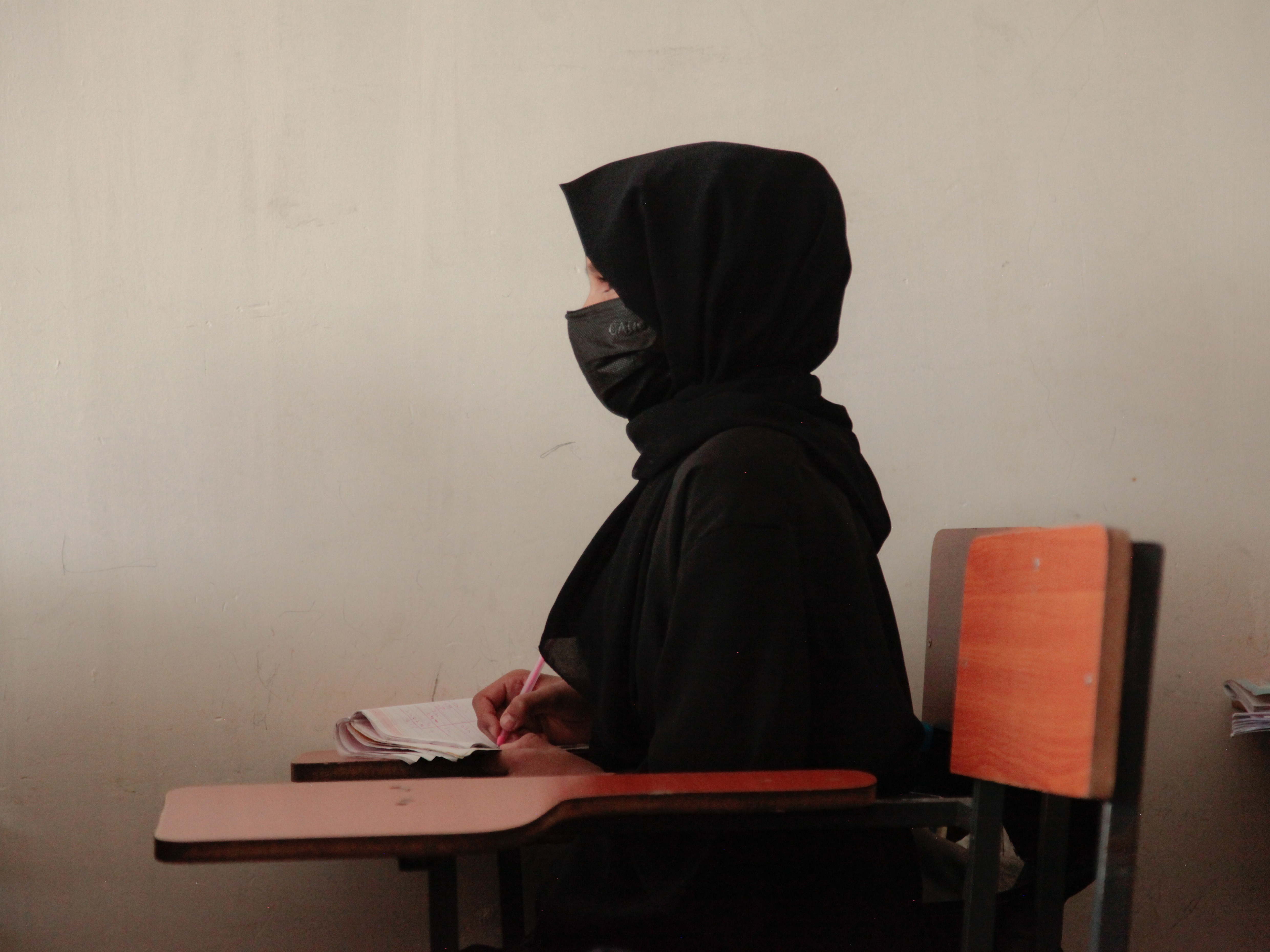 caption: A teenage girl wearing a face mask, head scarf and long black robe, listens to a math teacher at a tutoring center in Kabul. The center was established by a women's rights activist to circumvent a Taliban ban on girls attending secondary school. The activist said she has informal permission by Taliban authorities to run the center as long as teenage girls abide by a strict dress code.