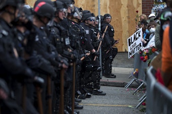 caption: Seattle police officers dressed in riot gear, left, stand in a line across 11th Street, opposite thousands of people protesting police brutality and the unjust deaths of Black Americans at the hands of law enforcement on the fifth day of protests following the violent police killing of George Floyd on Tuesday, June 2, 2020, in the Capitol Hill neighborhood of Seattle. 