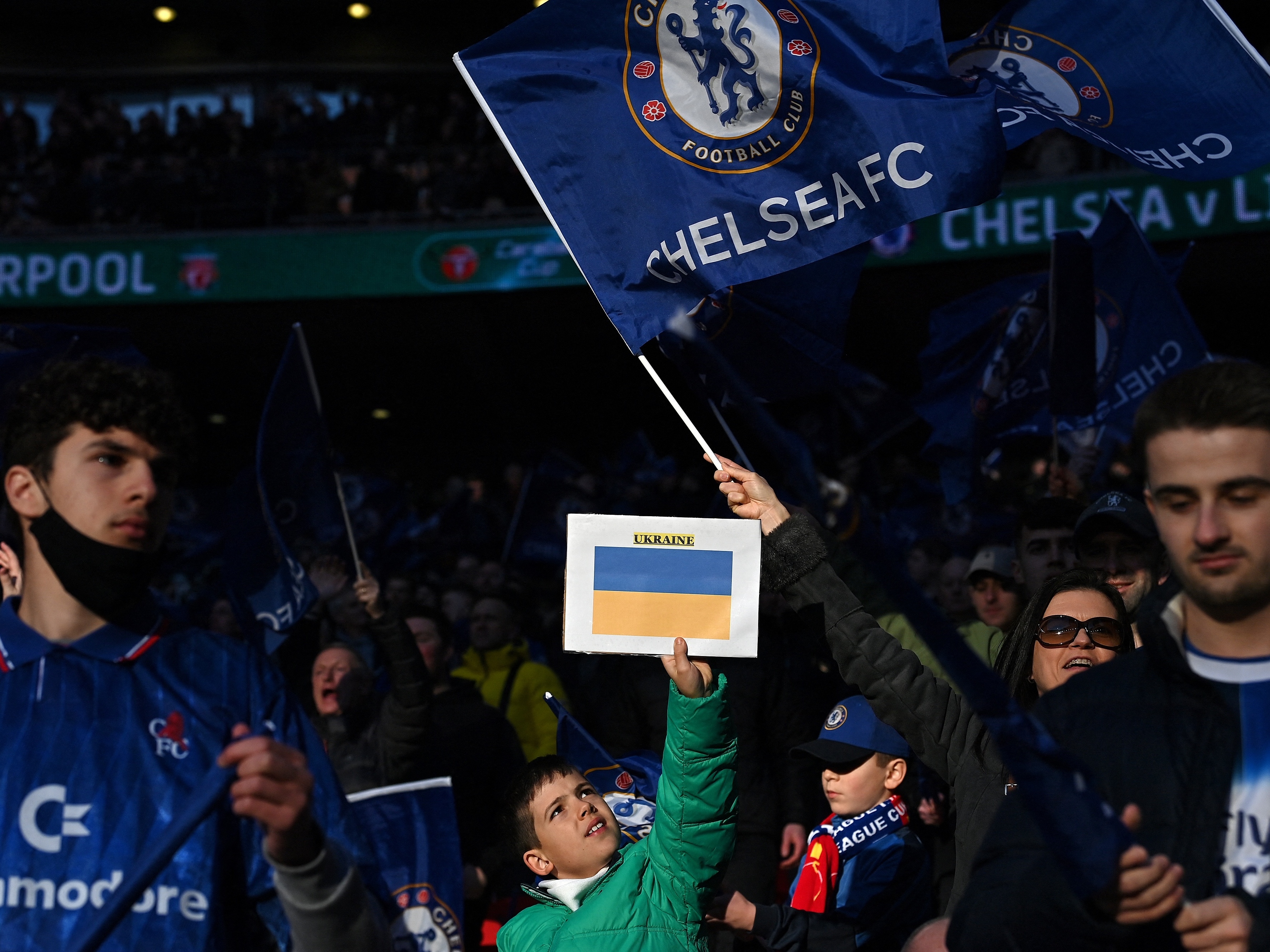 caption: Chelsea supporters hold up Ukrainian flags in the crowd ahead of the English League Cup final football match between Chelsea and Liverpool at Wembley Stadium, north-west London on February 27, 2022. Russian billionaire Roman Abramovich handed over control of Chelsea to trustees of its charitable foundation on Saturday as Premier League players showed their support for war-torn Ukraine.