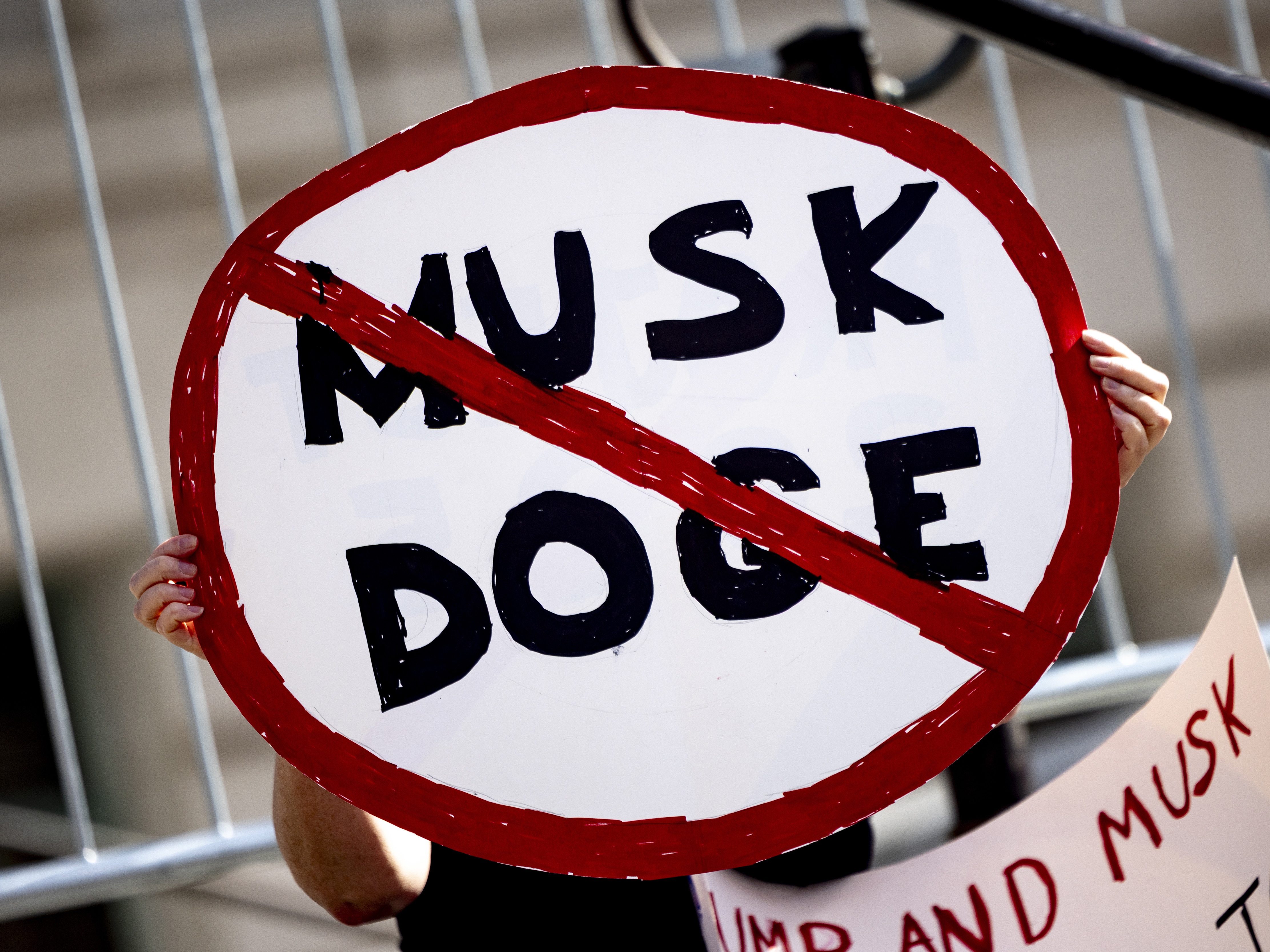 caption: A demonstrator holds a sign during a February rally in Los Angeles to protest President Trump's billionaire adviser Elon Musk and his DOGE team.